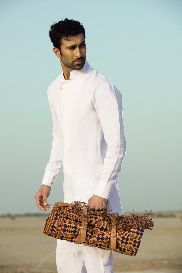 Man in white traditional outfit holding a patterned bag against a clear sky.