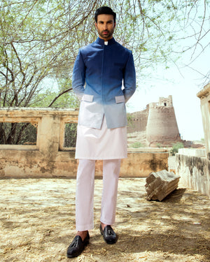 Man in a traditional outfit standing outdoors with trees and stone architecture in the background