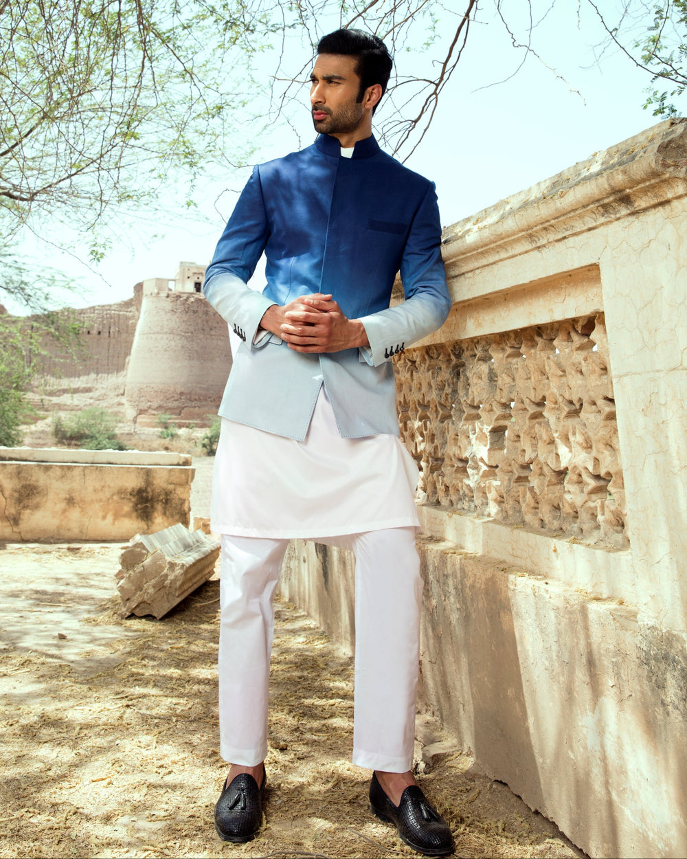 Man in a traditional outfit standing outdoors with trees and stone architecture in the background