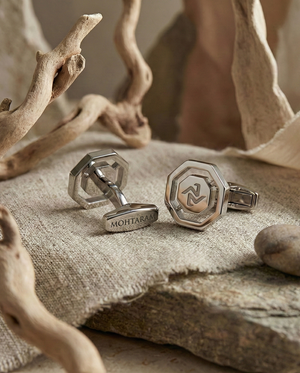 Silver cufflinks with a logo on a textured surface with natural elements