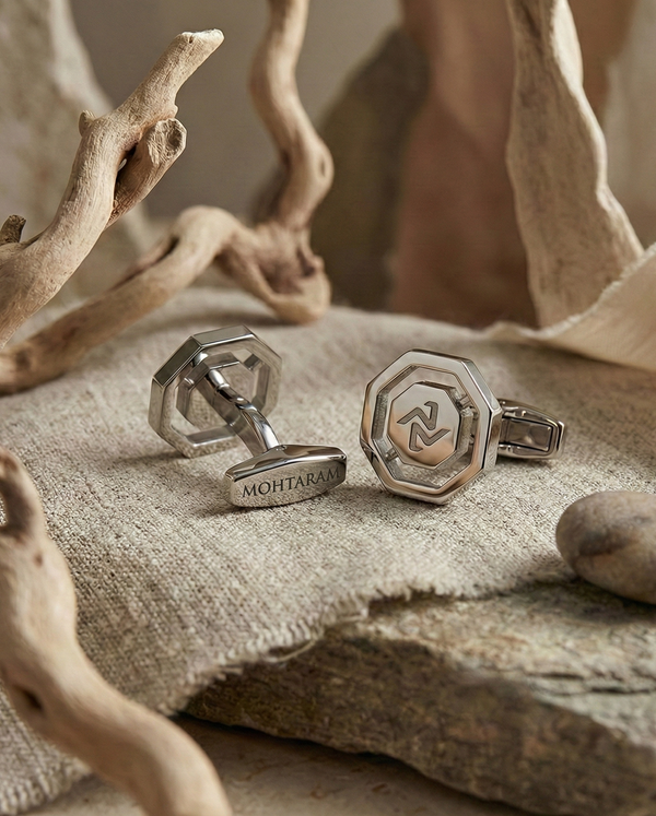 Silver cufflinks with a logo on a textured surface with natural elements