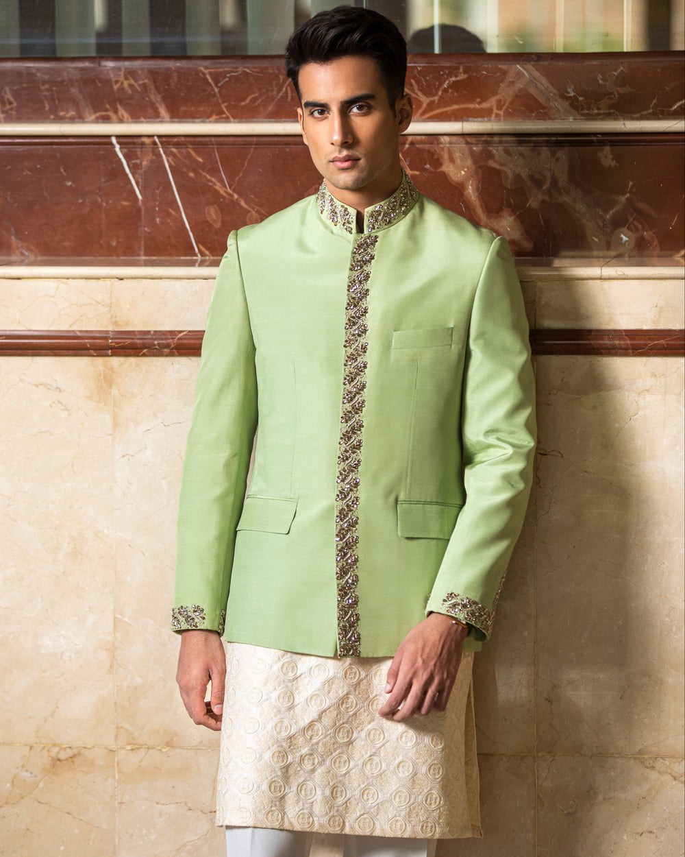 Man wearing a light green sherwani with white pants against a marble wall.