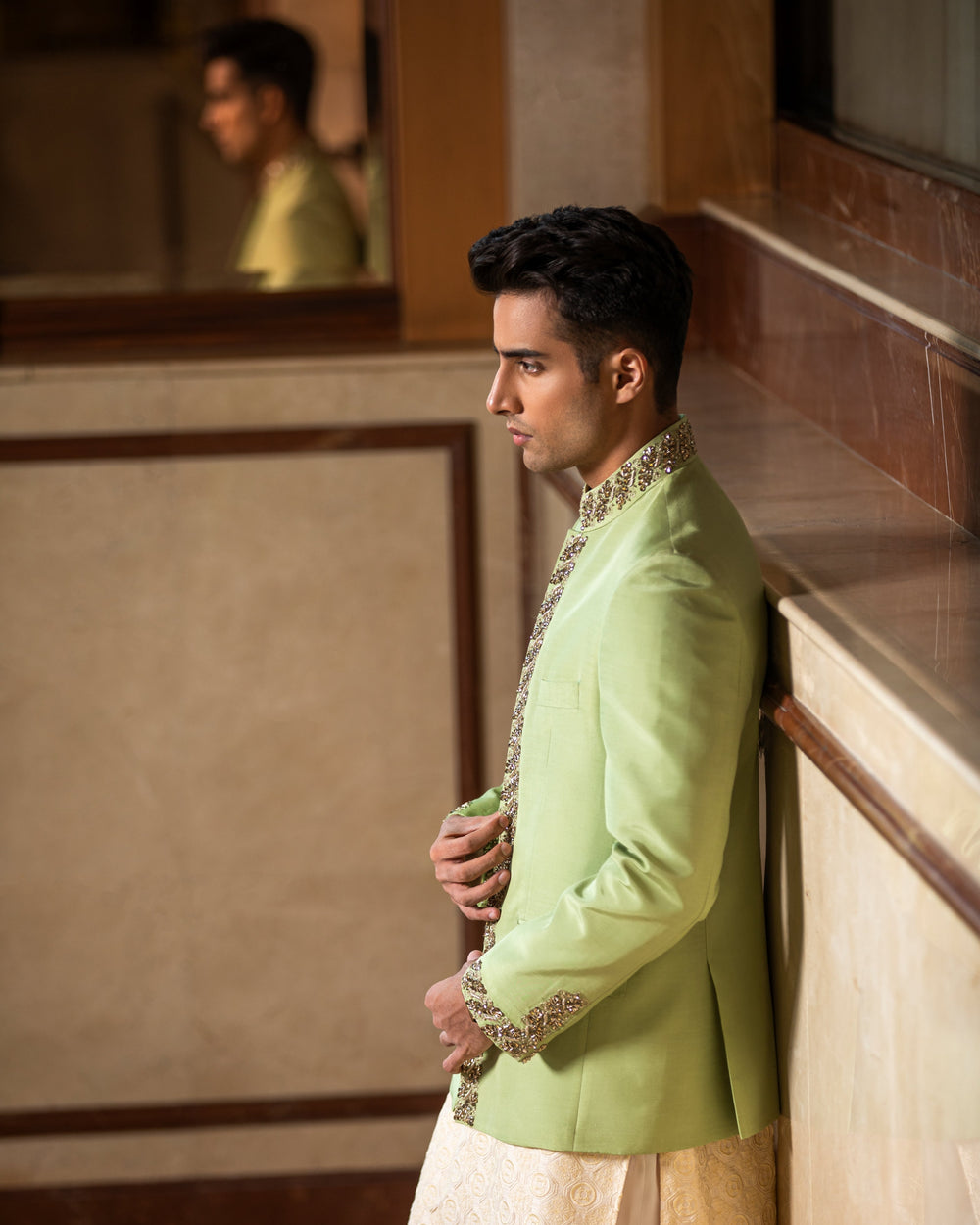 Man wearing a light green sherwani with white pants against a marble wall.