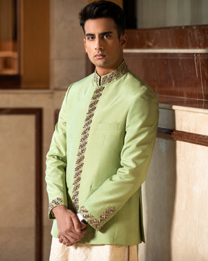 Man wearing a light green traditional outfit with intricate patterns in a room with marble walls.