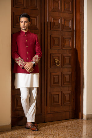 Man wearing a red sherwani with white pants standing in front of a wooden door.