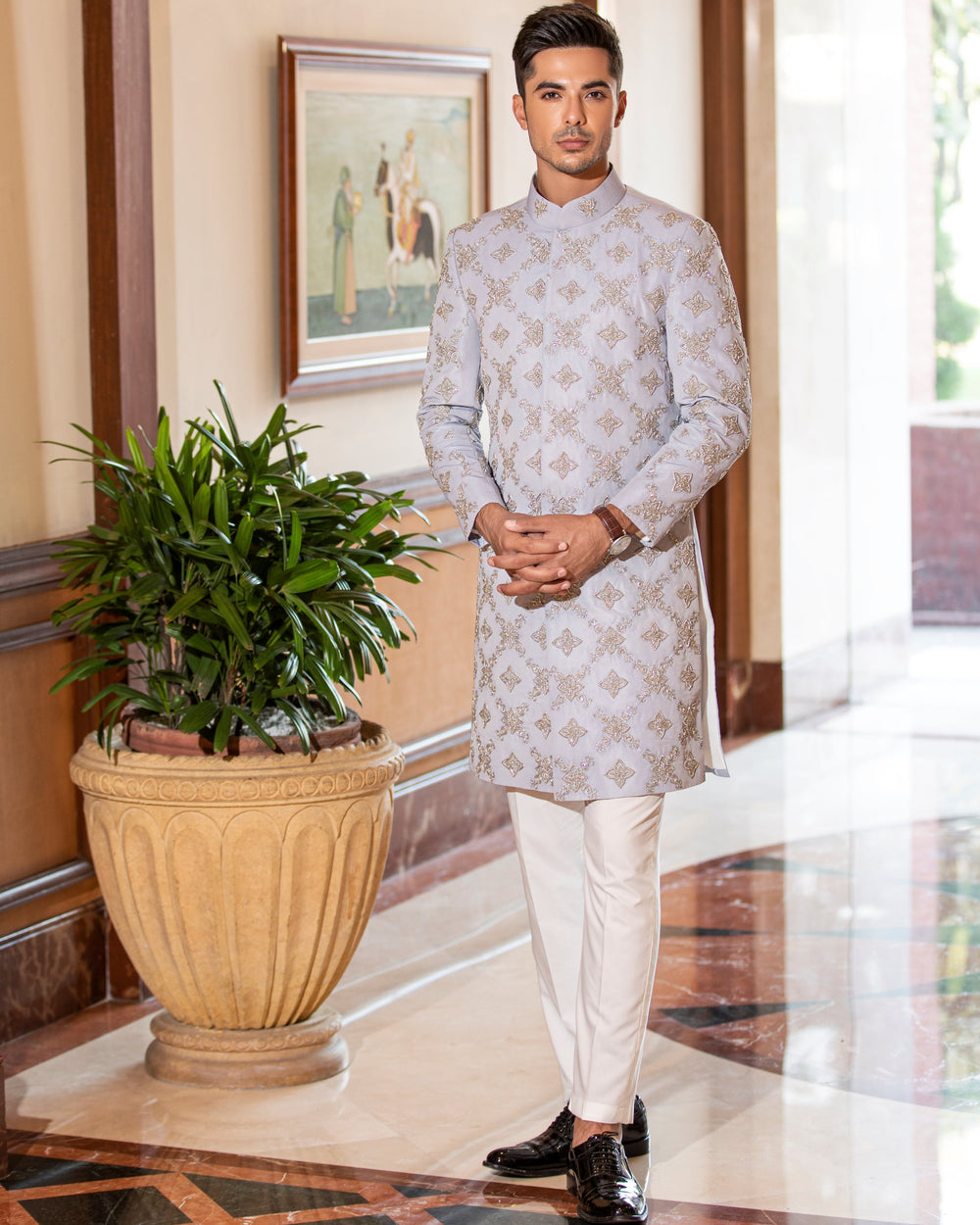 Man in traditional outfit standing in a well-decorated room with a plant and artwork.