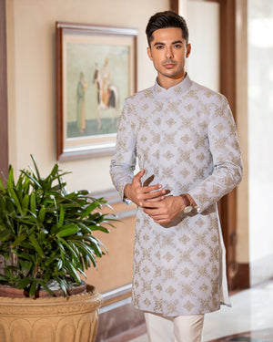 Man in a light gray traditional outfit standing indoors with a plant and framed picture in the background