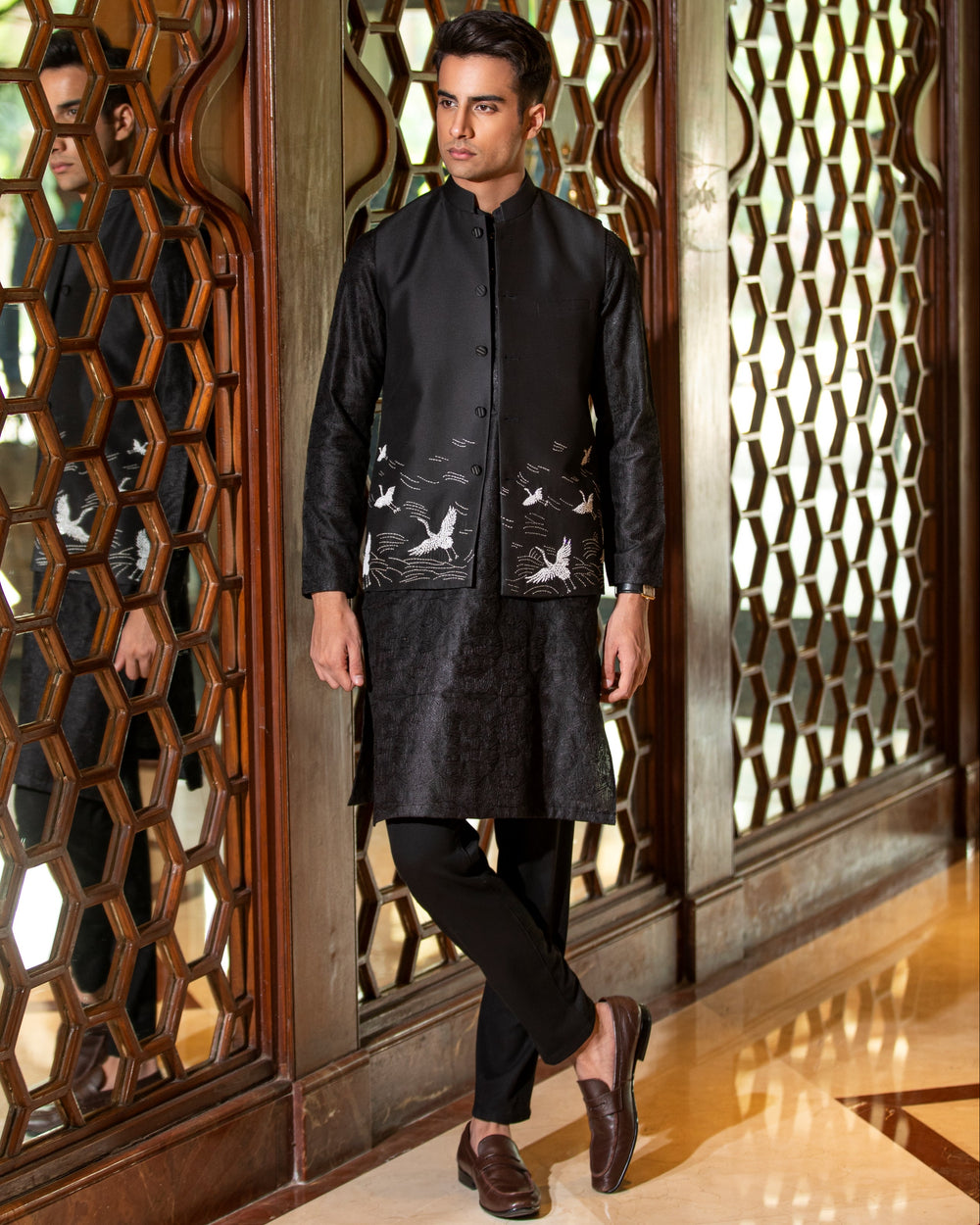 Man wearing a black traditional outfit with intricate patterns in a decorative setting.