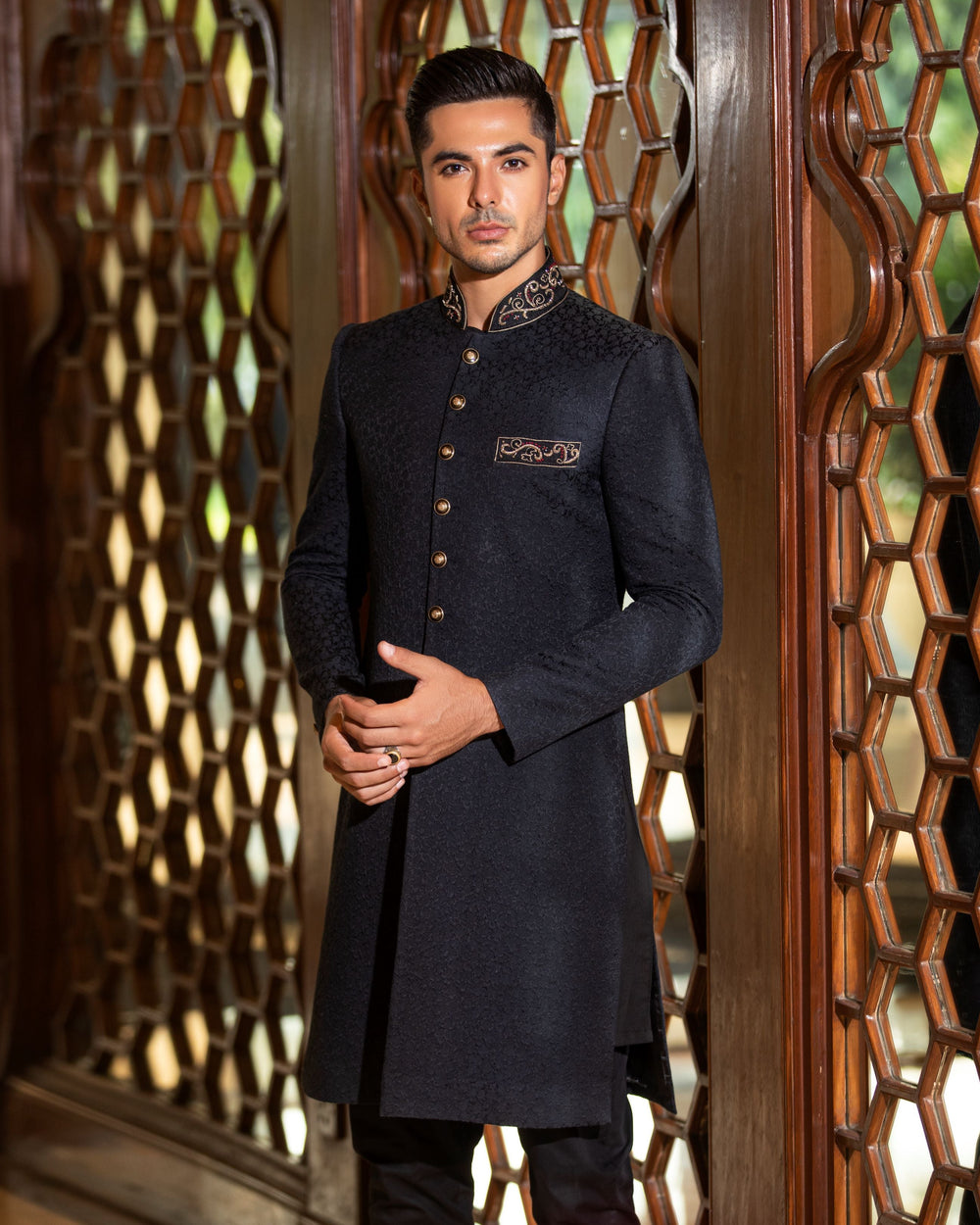 Man wearing a black traditional outfit standing in front of decorative wooden panels.