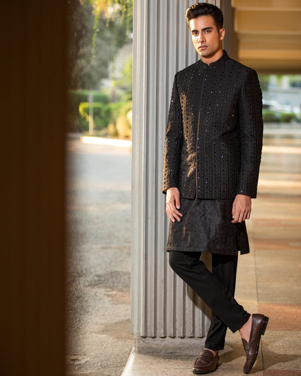 Man wearing a traditional black sherwani standing indoors with a blurred background