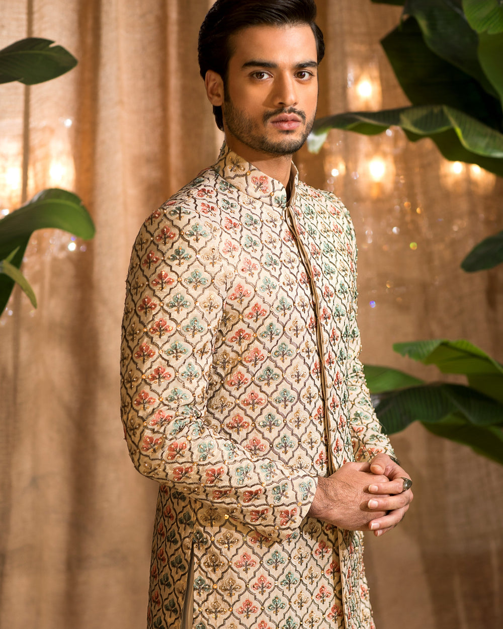 Man wearing a patterned traditional outfit with a beige curtain and green plants in the background