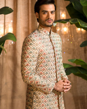 Man wearing a patterned traditional outfit with a beige curtain and green plants in the background