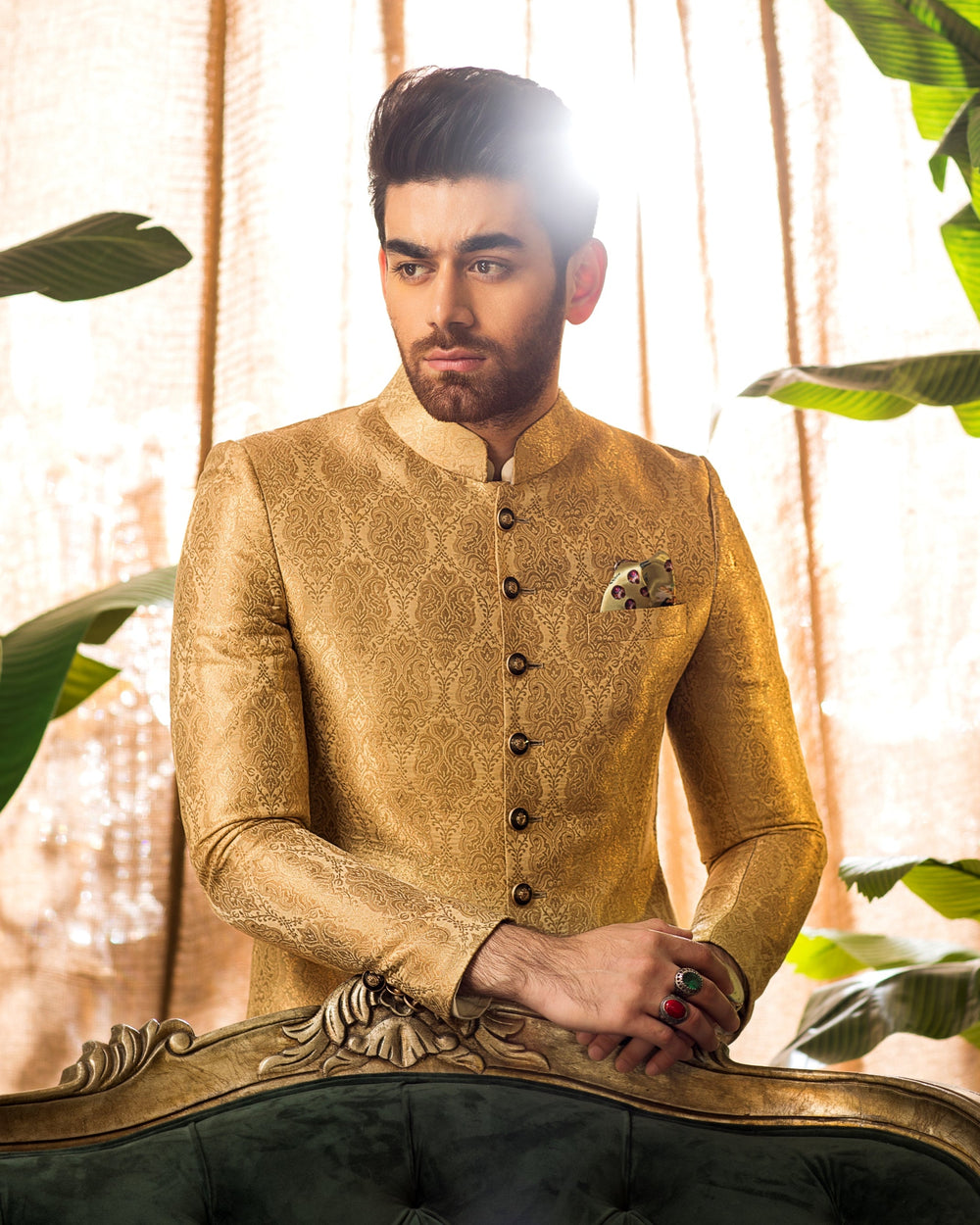 Man in a gold embroidered sherwani standing between large green leaves