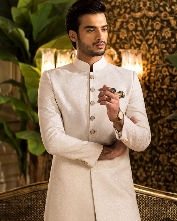 Man in a white sherwani standing in an elegant indoor setting with plants and decorative wall.