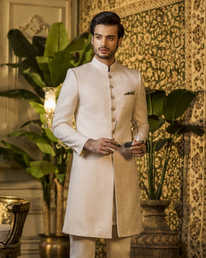 Man in a traditional cream sherwani standing in an ornate room with decorative elements.