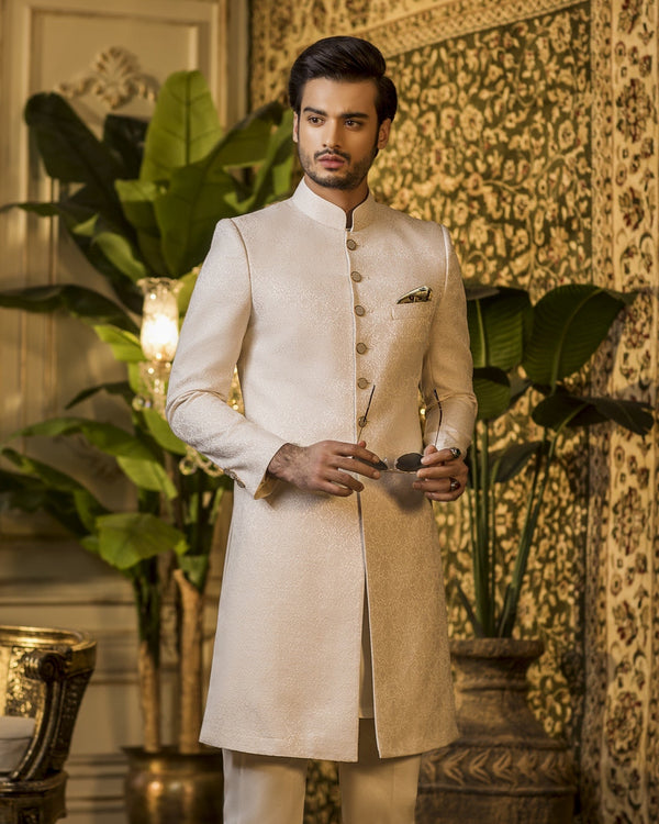 Man in a traditional cream sherwani standing in an ornate room with decorative elements.