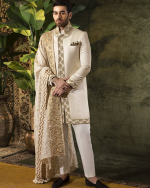 Man wearing a traditional sherwani with intricate patterns in a decorated indoor setting.