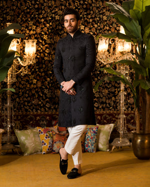 Man in a black kurta with white pants standing in a decorated room with plants and chandeliers.