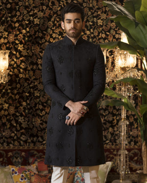 Man in a black kurta with white pants standing in a decorated room with plants and chandeliers.
