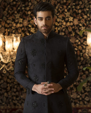 Man in a dark coat standing in front of a decorative wall with chandeliers.