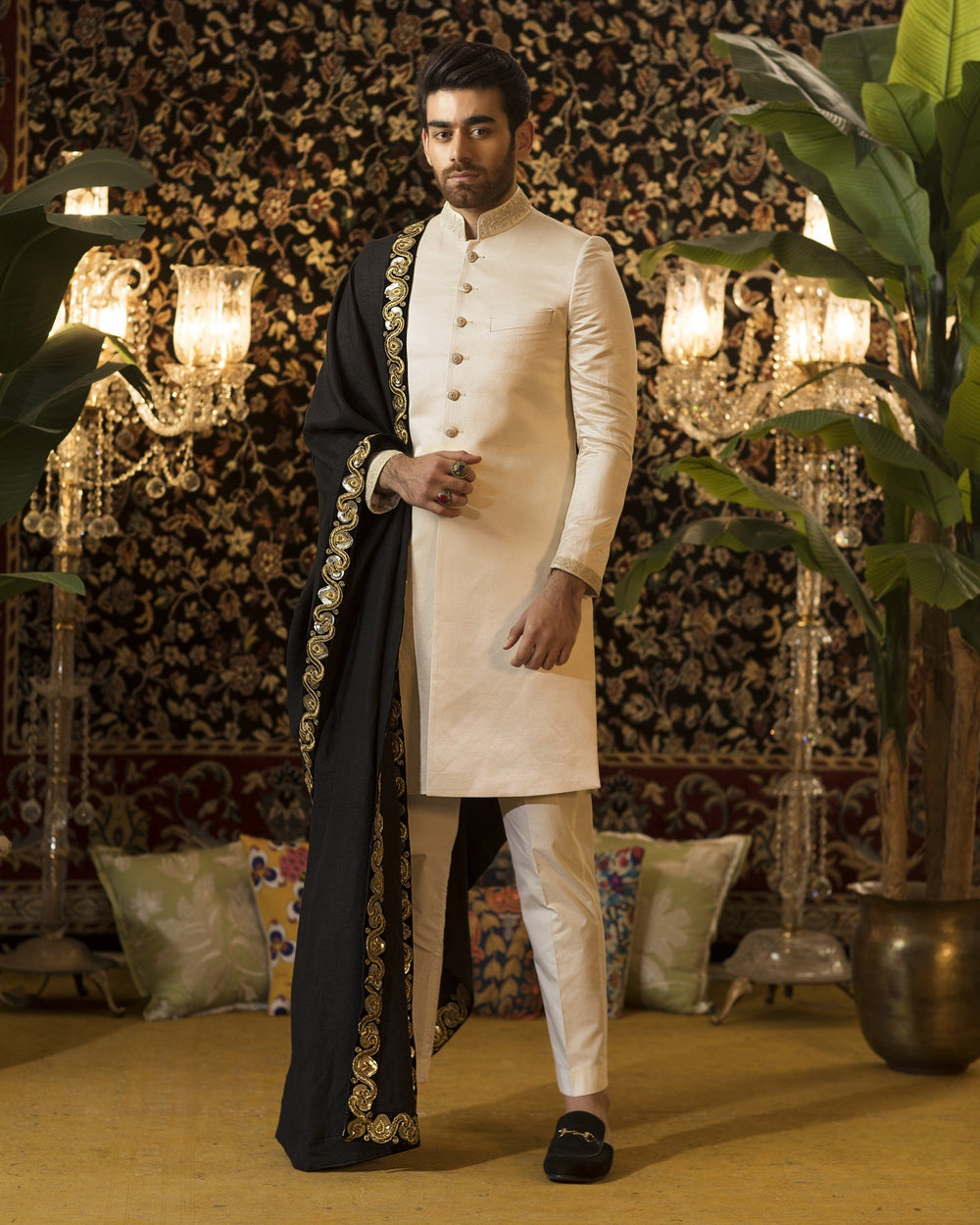 Man wearing a traditional outfit with gold embroidery against a decorative wall.