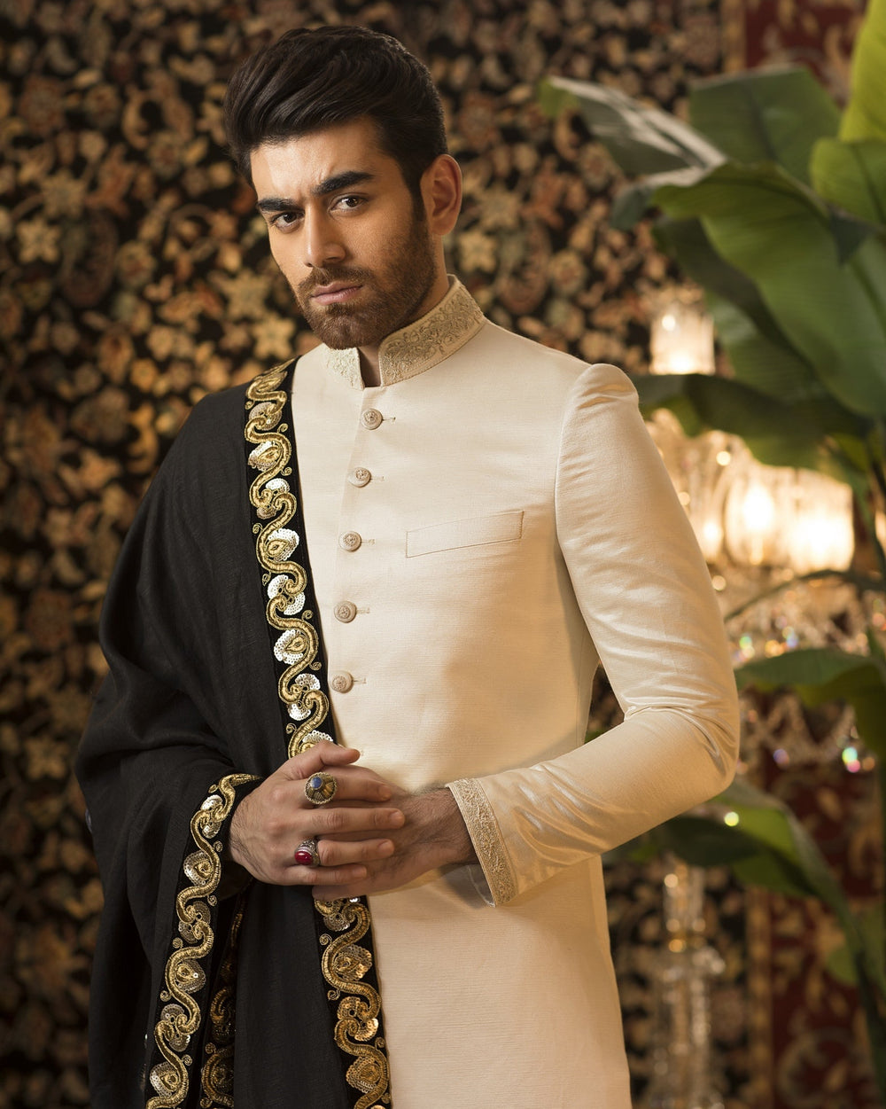 Man wearing a traditional outfit with gold embroidery against a decorative wall.