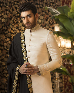 Man wearing a traditional outfit with gold embroidery against a decorative wall.