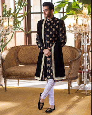 Man in a black sherwani standing in an elegant room with decorative elements.