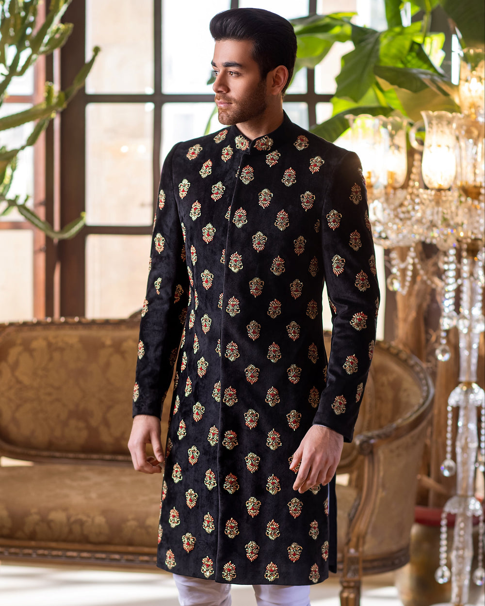 Man in a black sherwani standing in an elegant room with decorative elements.