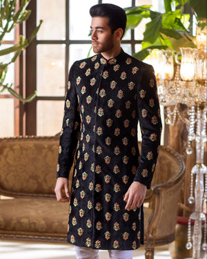 Man wearing a black and gold patterned kurta with white pants in an elegant room.