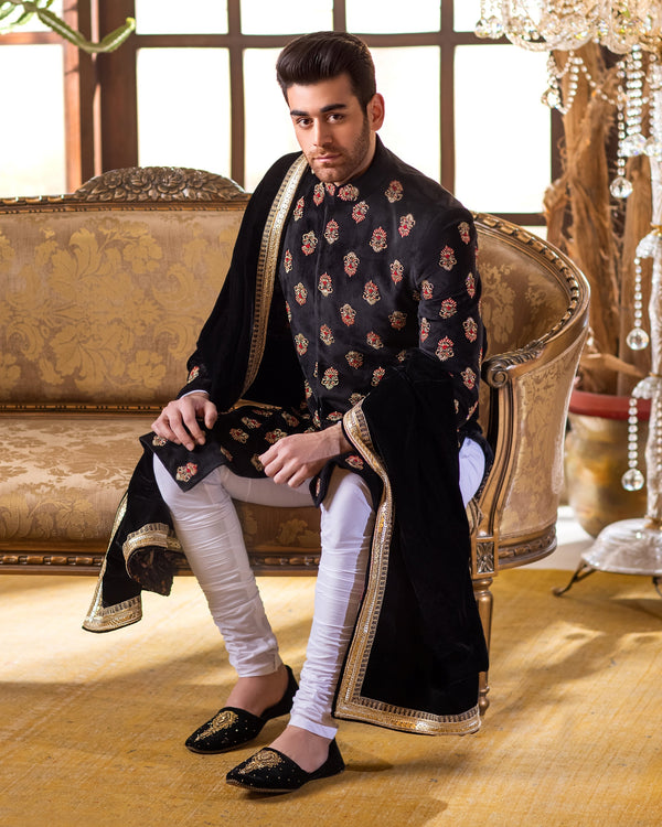 Man in traditional attire sitting on an ornate couch with decorative plants and chandeliers in the background.