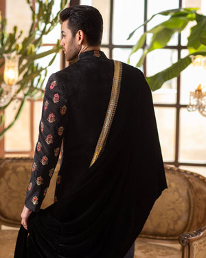 Man wearing a black traditional outfit with intricate patterns, standing in a room with large windows and plants.