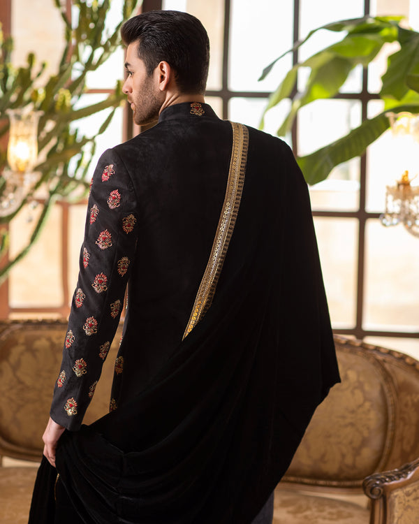 Man wearing a black traditional outfit with intricate patterns, standing in a room with large windows and plants.