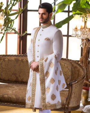 Man wearing a traditional sherwani in a decorated room with plants and furniture.
