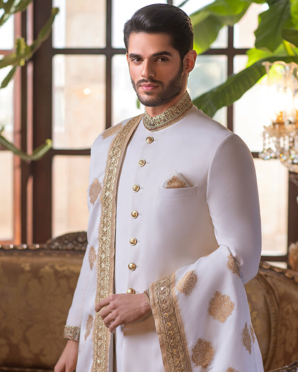Man in a white sherwani with gold embroidery standing in an elegant room with chandeliers and a couch.