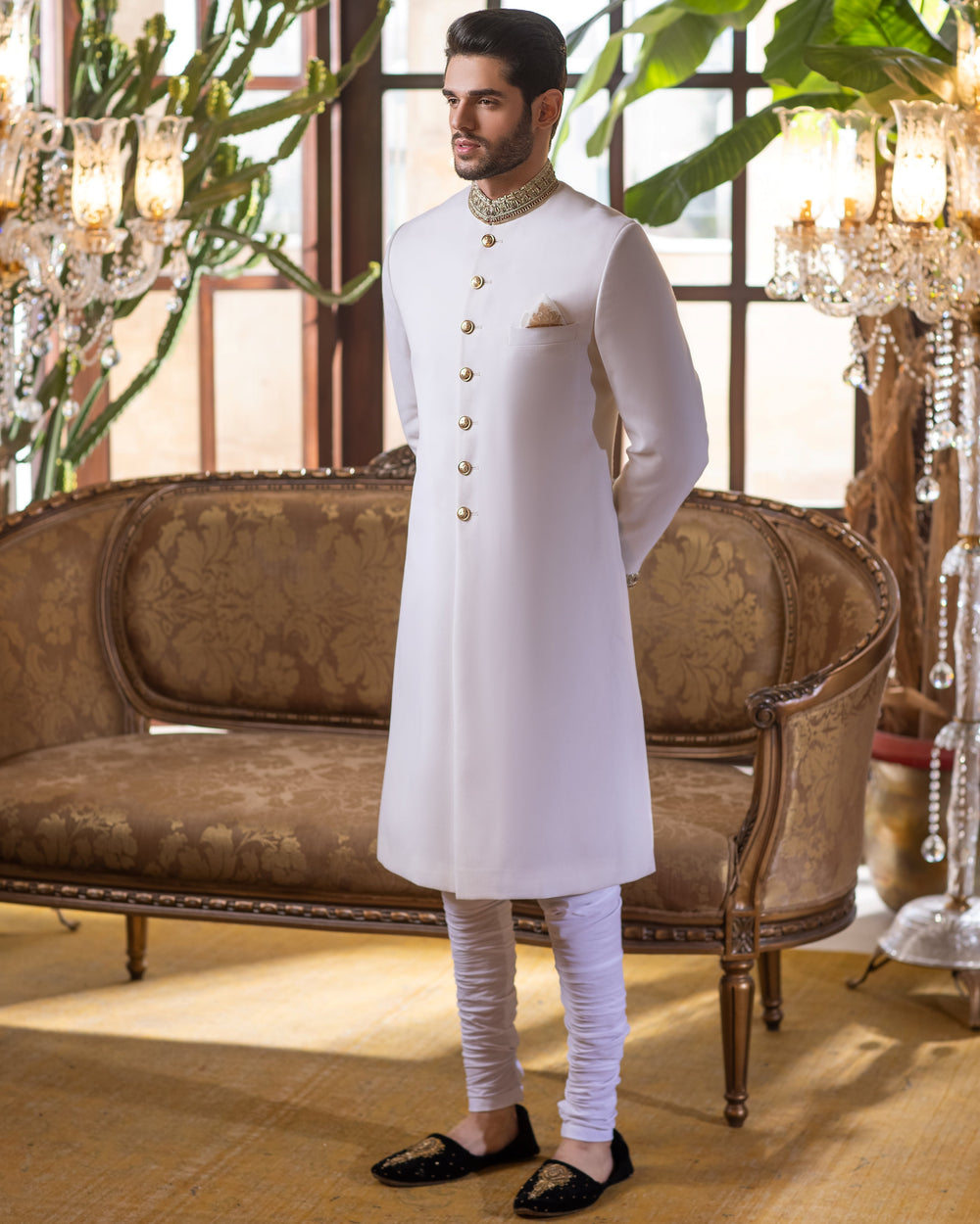 Man in a white sherwani with gold embroidery standing in an elegant room with chandeliers and a couch.