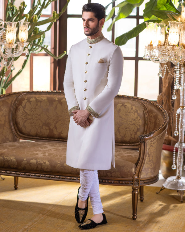 Man in a white sherwani standing in an elegant room with chandeliers and a sofa.