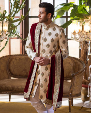 Man in traditional sherwani standing in an elegant indoor setting with chandeliers and chairs.