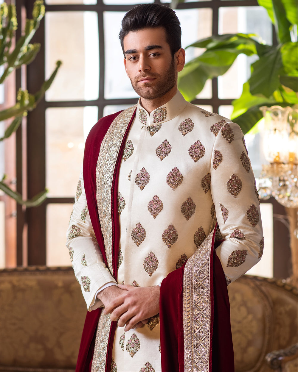 Man in a traditional outfit standing in an elegant room with chandeliers and plants.