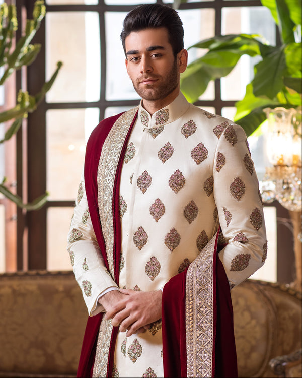 Man in a traditional outfit standing in an elegant room with chandeliers and plants.