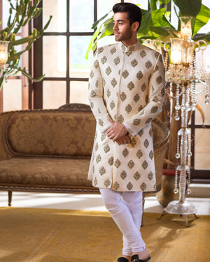 Man in a traditional white sherwani with gold patterns standing in an elegant room.