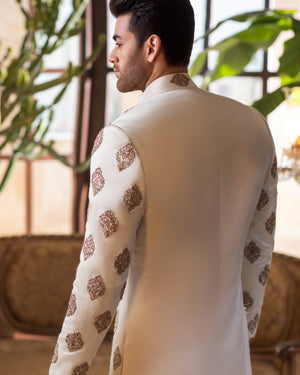 Man wearing a traditional outfit with intricate patterns in a room with plants and furniture.