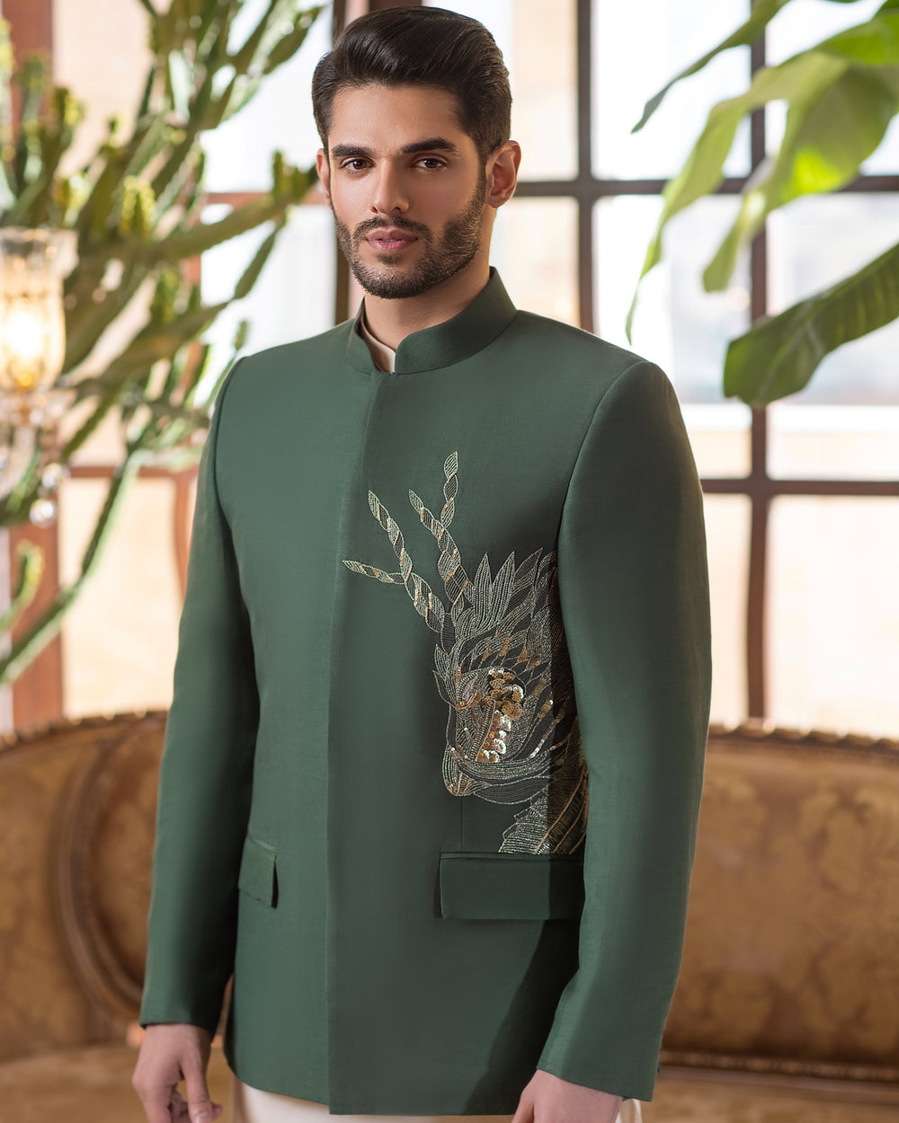 Man in a green kurta with white pants standing in a stylish room with plants and furniture.