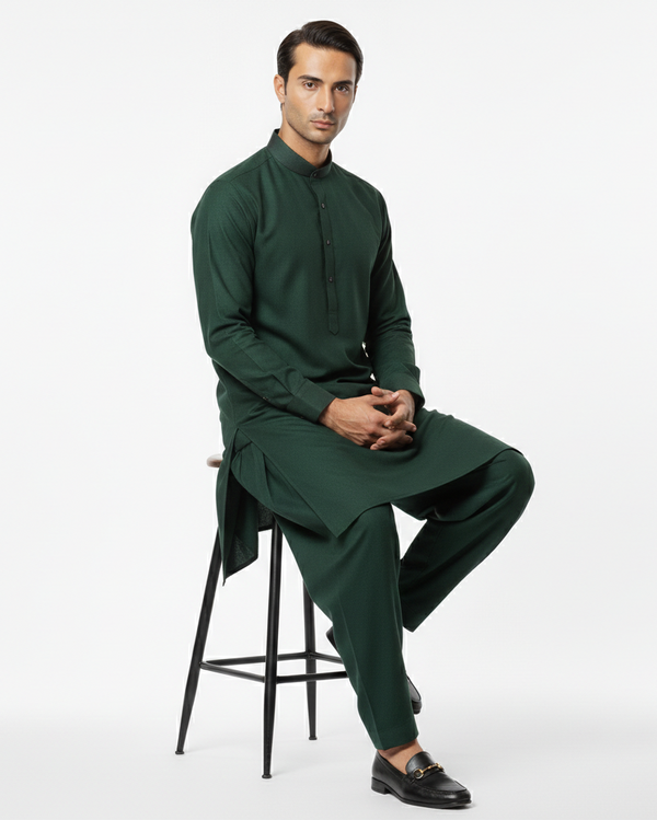 Man wearing a dark green traditional outfit sitting on a stool against a white background