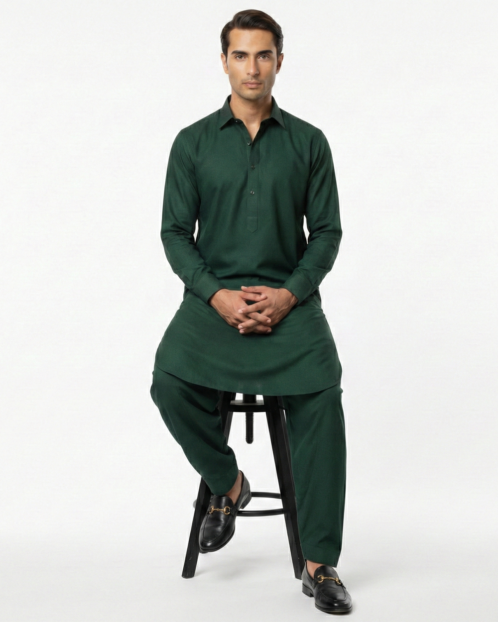 Man wearing a dark green traditional outfit sitting on a stool against a white background