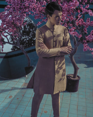 Man in a beige suit standing next to a potted tree with pink blossoms on a tiled floor.
