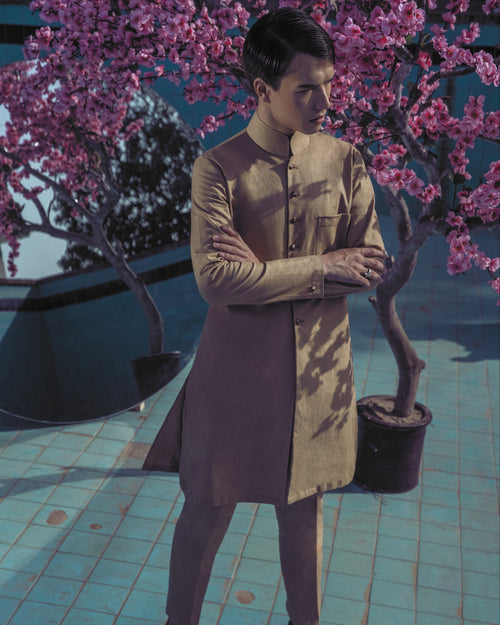 Man in a beige suit standing next to a potted tree with pink blossoms on a tiled floor.