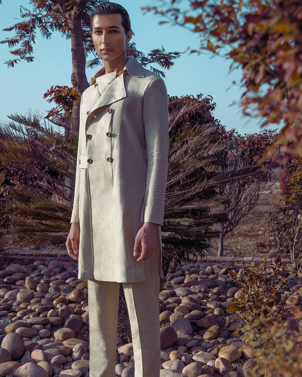 Man in a light-colored suit standing on a rocky path with trees and shrubs in the background