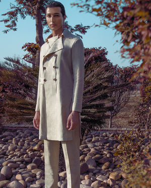 Man in a light-colored suit standing on a rocky path with trees and shrubs in the background