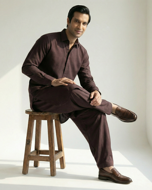 Man in a brown outfit sitting on a wooden stool in a bright room.